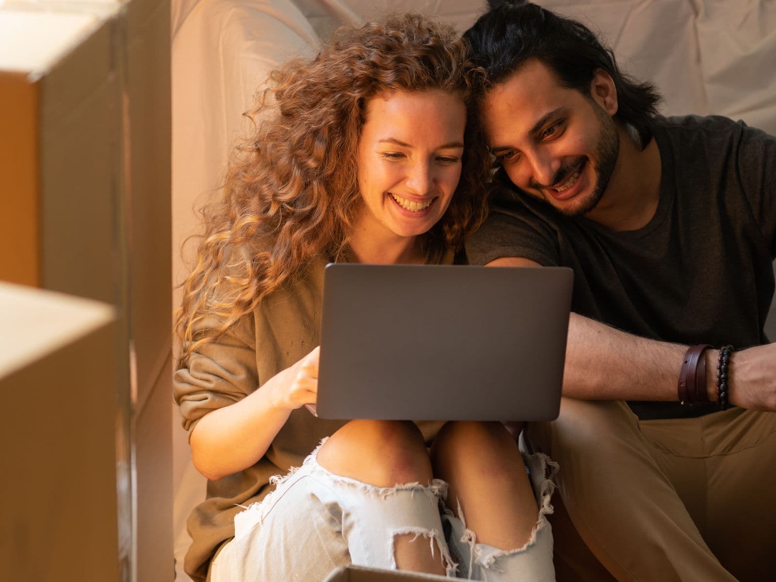 A couple settling into a new home, smiling as they plan together on a laptop among moving boxes